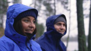 En ung kvinne og mann sitter ute i skogen, ser framfor seg og smiler. De har blå regnjakker og hette på. Brukt i forbindelse med Roza Pixel blogginnlegg. Bilde tatt ifbm reklame for Jotunheim og Sport 1.