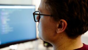 Nærbilde av bakhodet av mann med briller som ser på dataskjerm med koding. Fra Cappelen Damm rekrutteringsfilm av Roza Pixel, videoproduksjon.