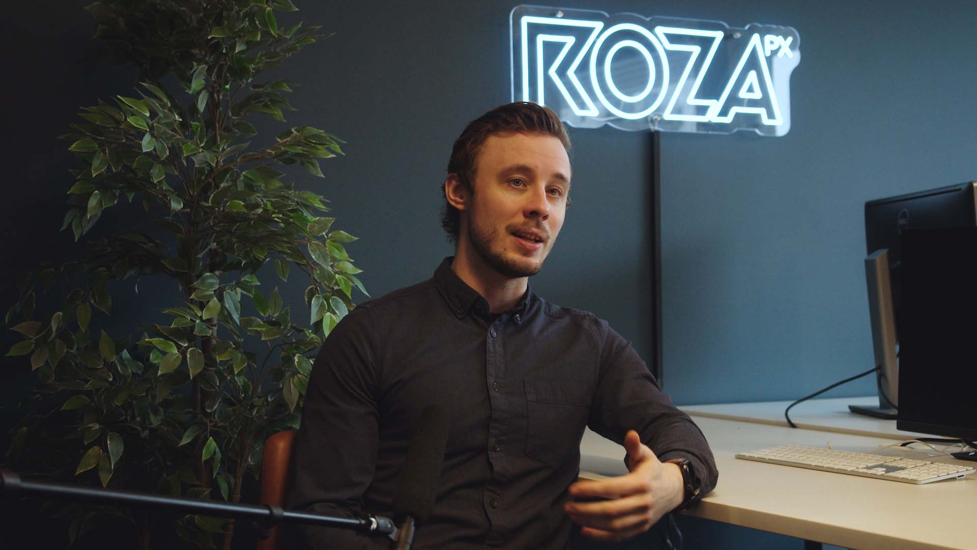 En ung mann sitter i et kontor eller podcast-studio og prater. Blå vegg bak han, Roza logo på veggen. Brukt i forbindelse med bloggpost hos Roza om å bli komfortabel foran kamera.
