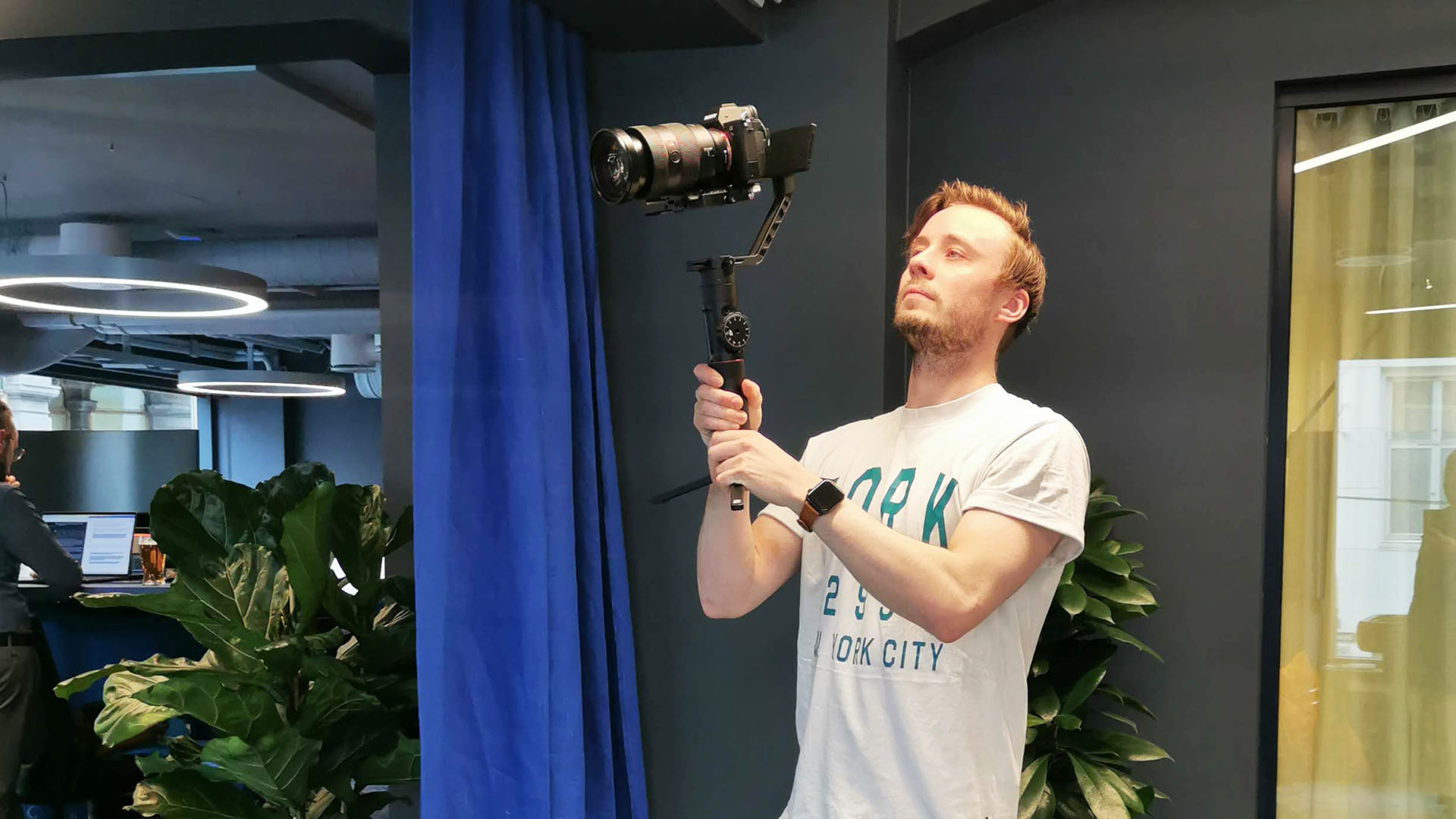 En ung mann står på et blått kontor med et videokamera på en gimbal stabilisator over hodet. Brukt i forbindelse med blogginnlegg hos Roza Pixel om videoproduksjon og tabber.